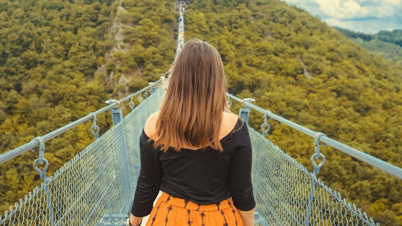 Una mujer de espaldas que camina hacia adelante en un puente colgante largo y estrecho que se extiende sobre una montaña verde por las copas de los árboles. El cielo es claro con algunas nubes.